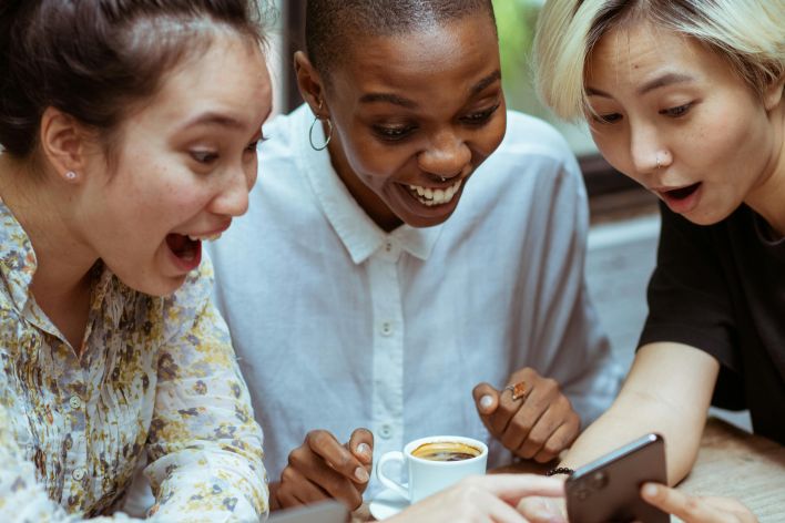 Amazed surprized multiethnic female friends in casual clothes with cup of coffee browsing smartphone while sitting at wooden table with laptop against window